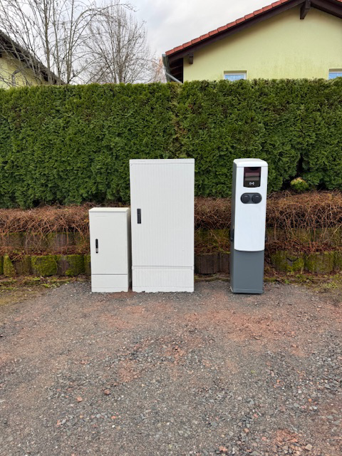 Verteilerschränke und Ladesäule an einem Stellplatz vor einer Hecke